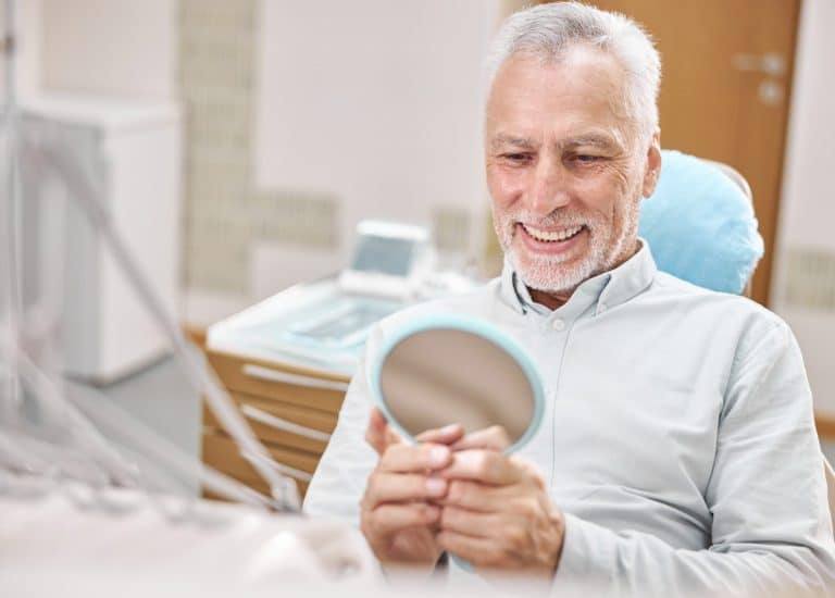 Un bărbat în vârstă zâmbind cu un implant dentar Straumann într-o oglindă la cabinetul stomatologic din Rahova, Sector 5.